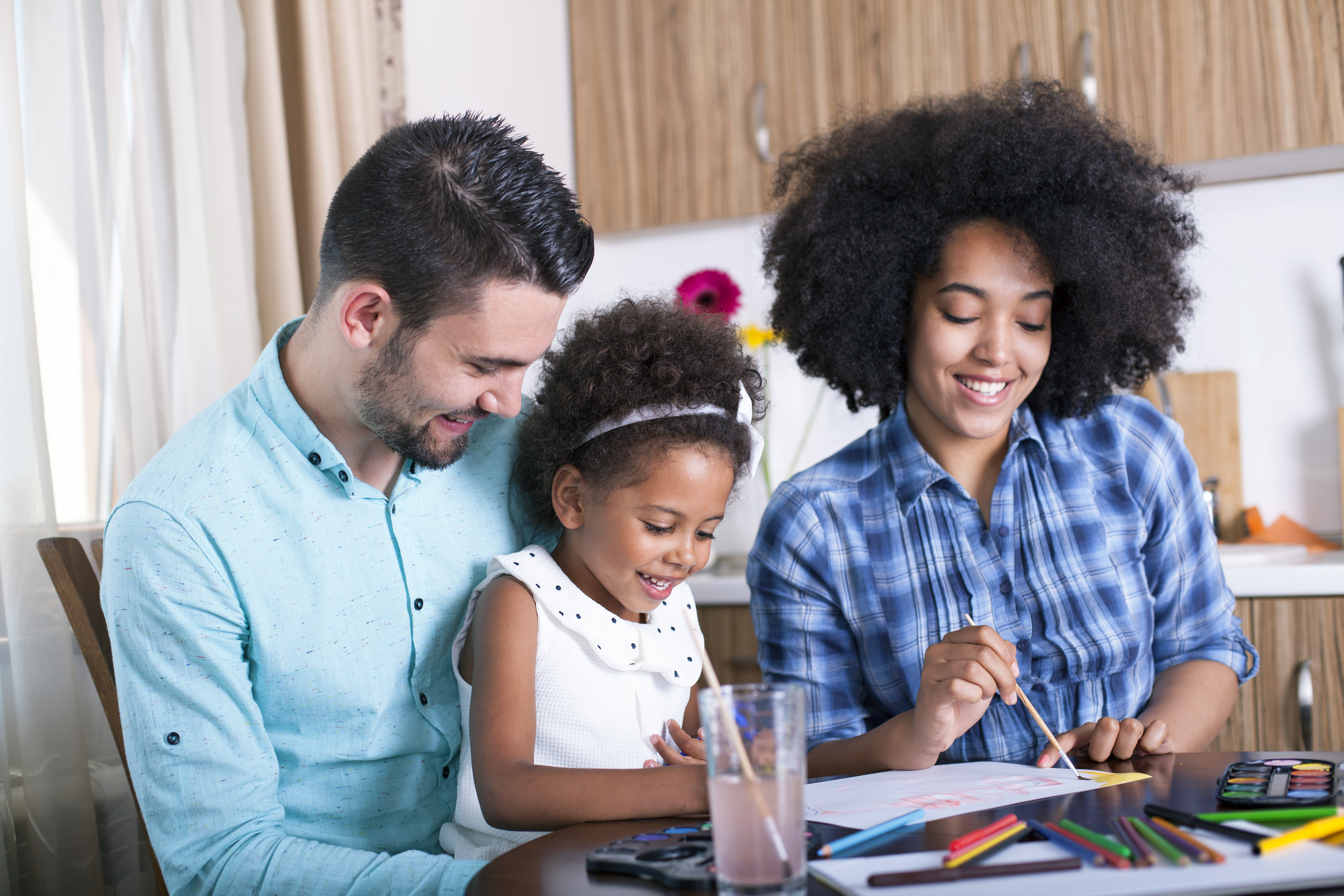 A smiling father, mother and daughter sit at a table doing crafts. The daughter is sitting on her father's lap.